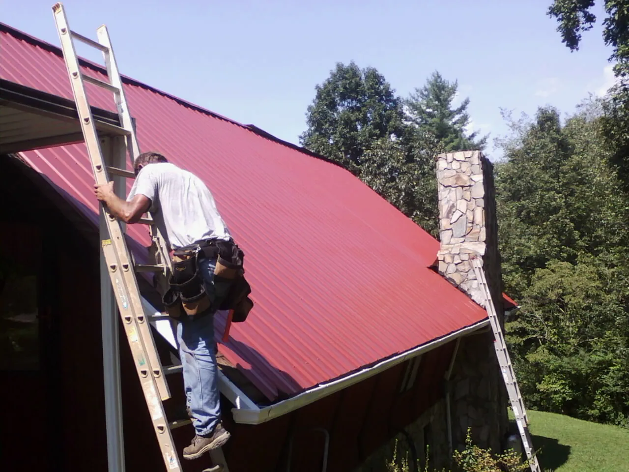Roofer on a ladder beside a red metal roof and stone chimney — Soffit & Fascia Repair by Roofing Brothers in Northbrook